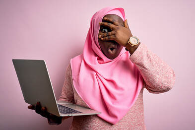 Woman in a pink headscarf holds laptop and peers out from behind her fingers