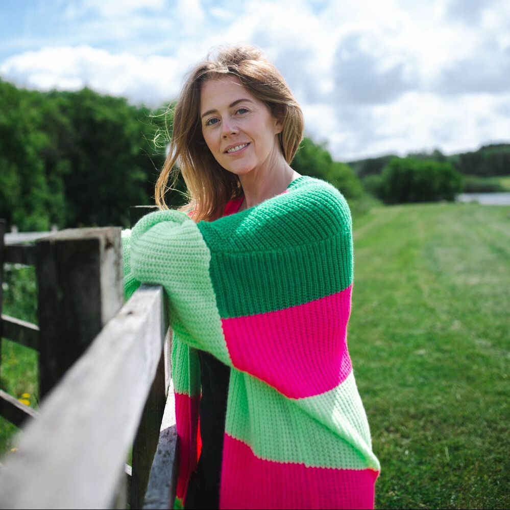 Clare wears a pink and green striped jumper and stands by a gate, smiling at the camera